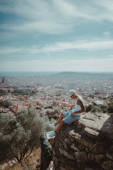 A man sits on a rock formation overlooking a panoramic view of Barcelona in summer.