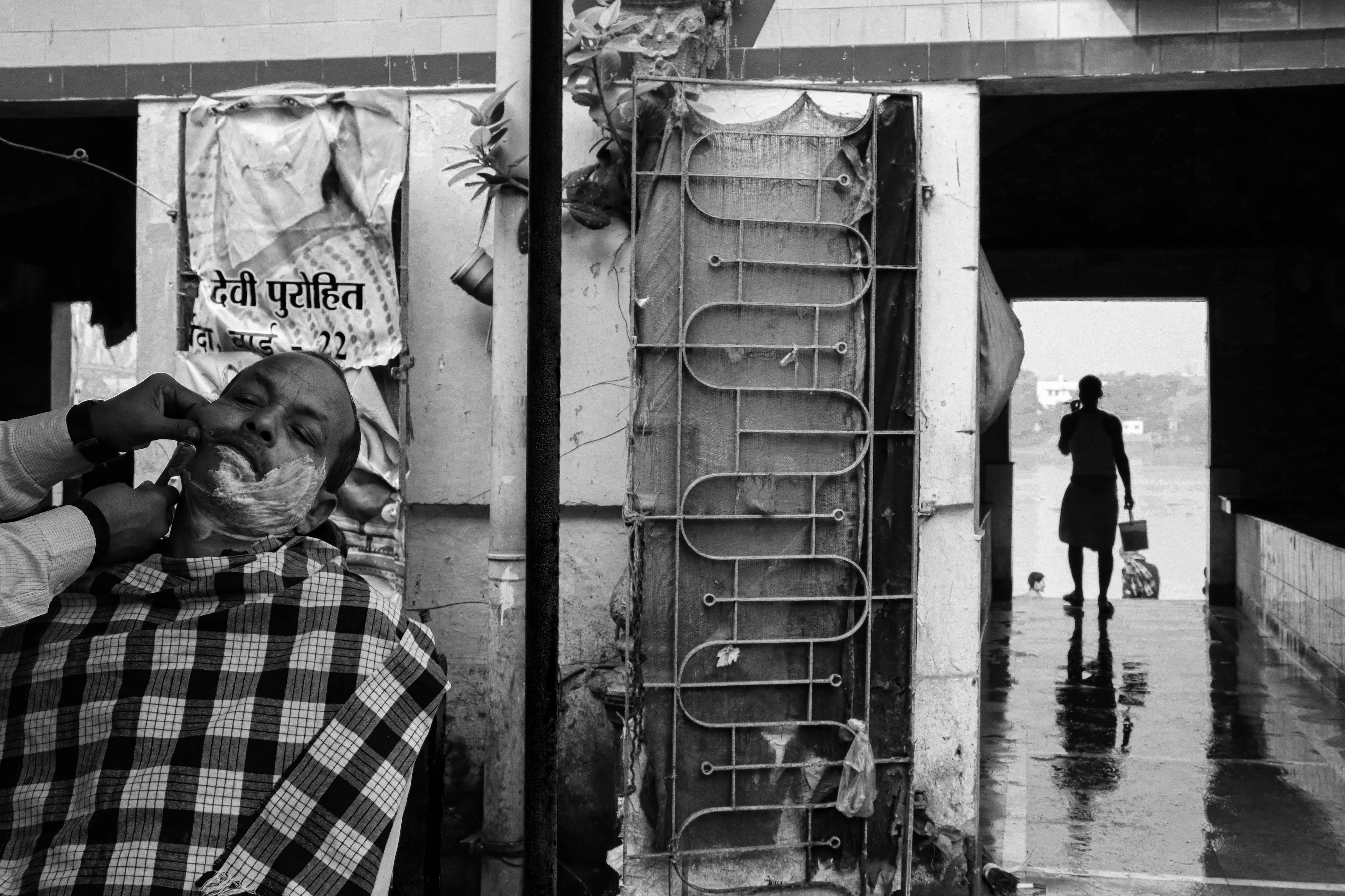 A street barber shaves a man in Kolkata, showcasing urban life. Black and white photography.