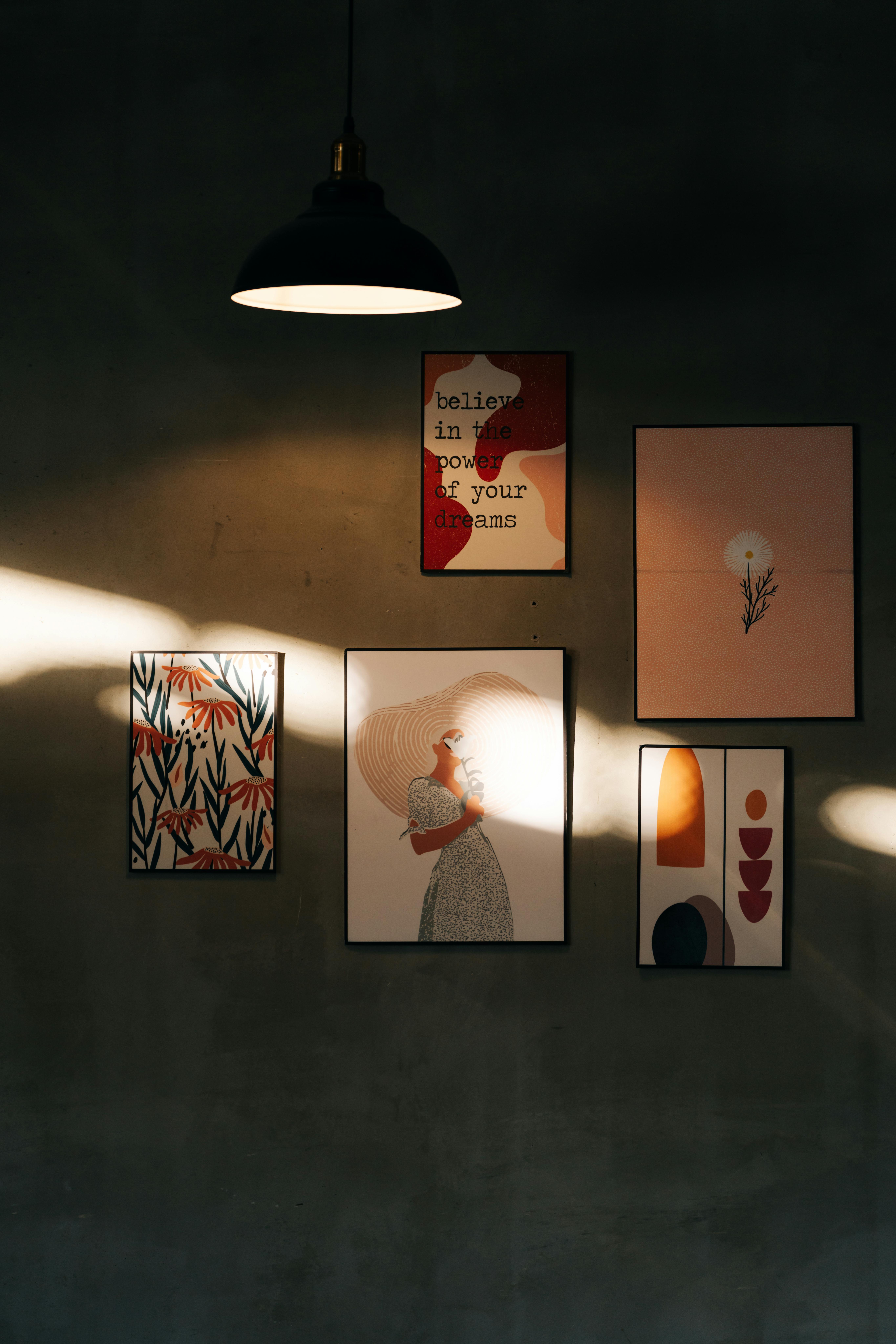 A collection of artistic wall frames illuminated by a sunbeam under a hanging lamp.