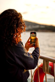 A woman taking a sunset photo over the sea from a boat, capturing the serene seaside view.