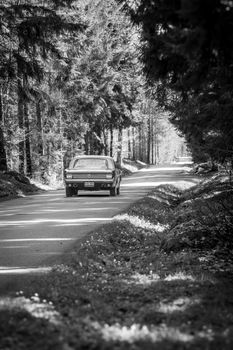 A classic vintage car travels down a secluded forest road, captured in black and white.