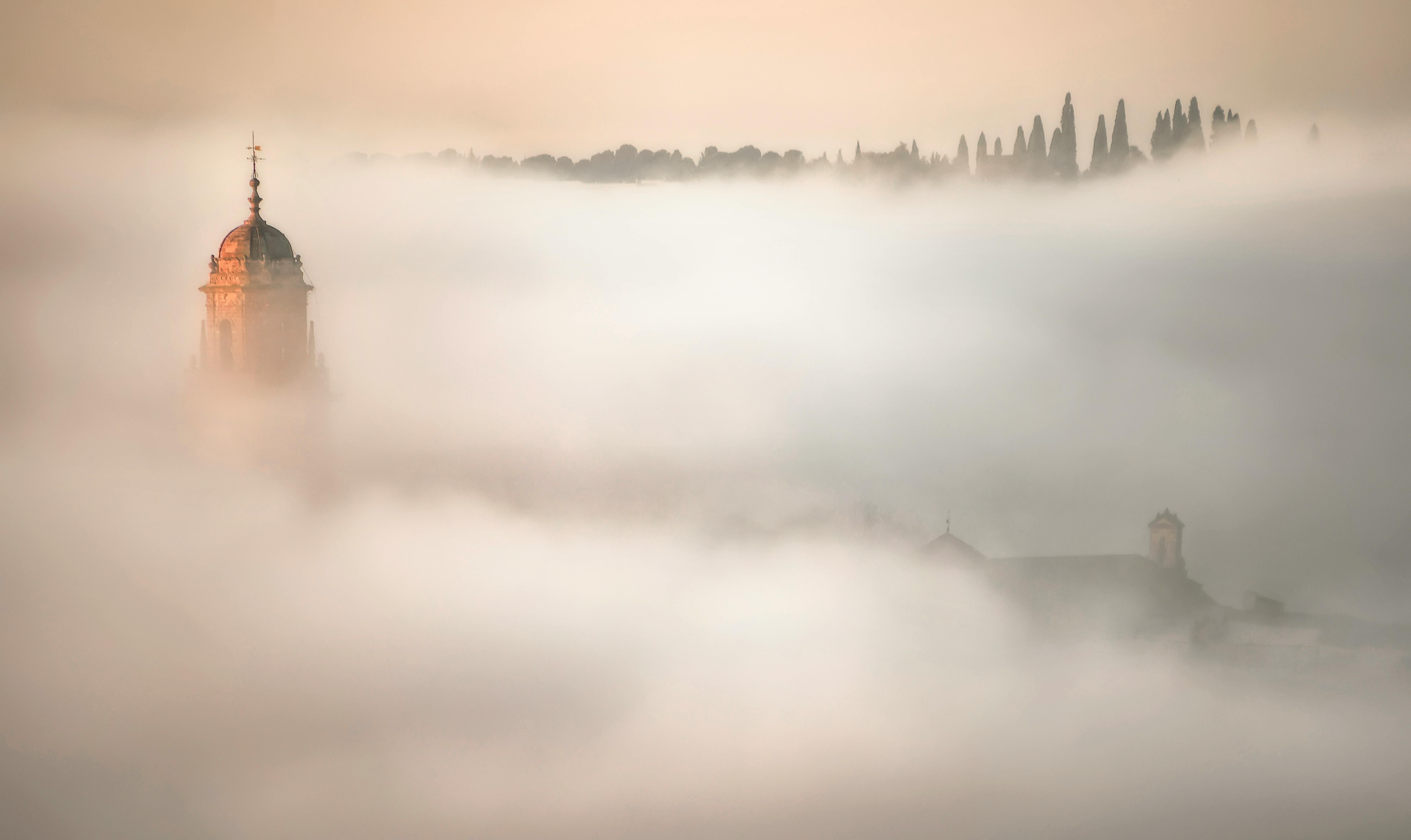 Captivating view of a church tower emerging from dense fog in Córdoba, Spain, with a serene forest backdrop.
