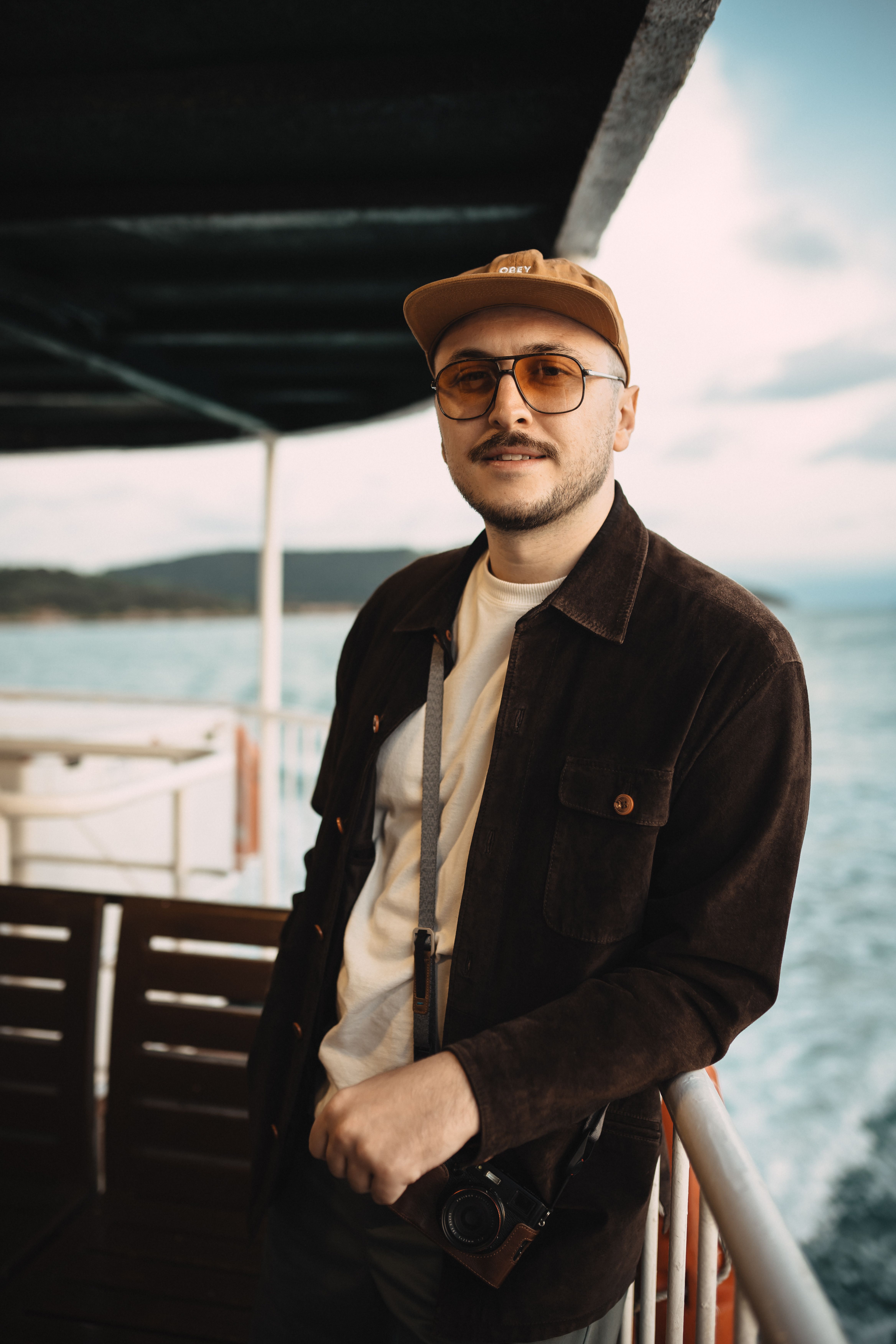 Man wearing cap and sunglasses casually posing on a boat deck with a scenic ocean backdrop.