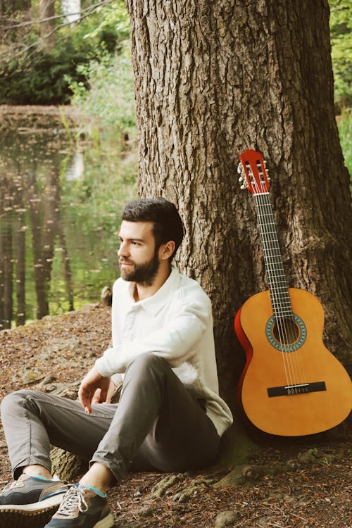 A man sitting next to a tree with an acoustic guitar