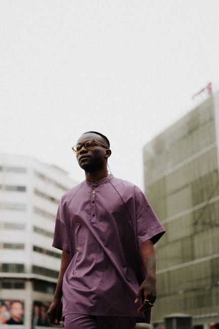 Man wearing glasses in a purple shirt, walking through an urban city street with modern buildings.