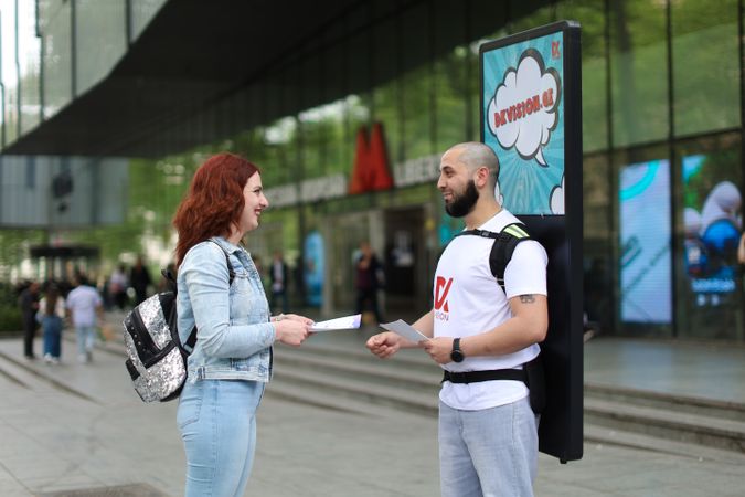 A man promoting with a backpack sign interacts with a woman on a busy city street.