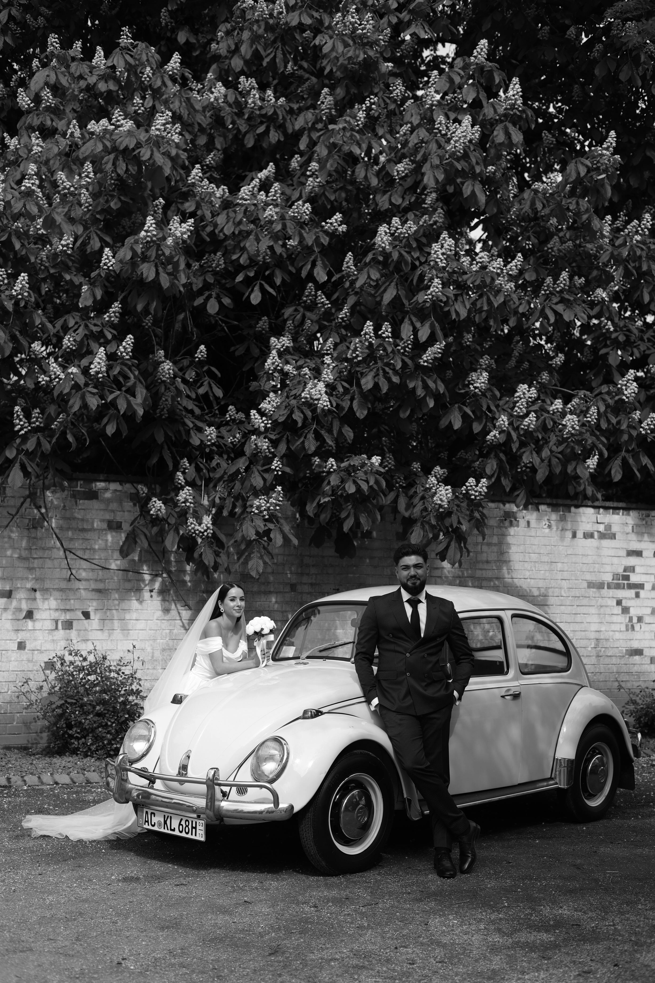 Elegant black and white wedding photo featuring a couple with a vintage car in a rural German setting.