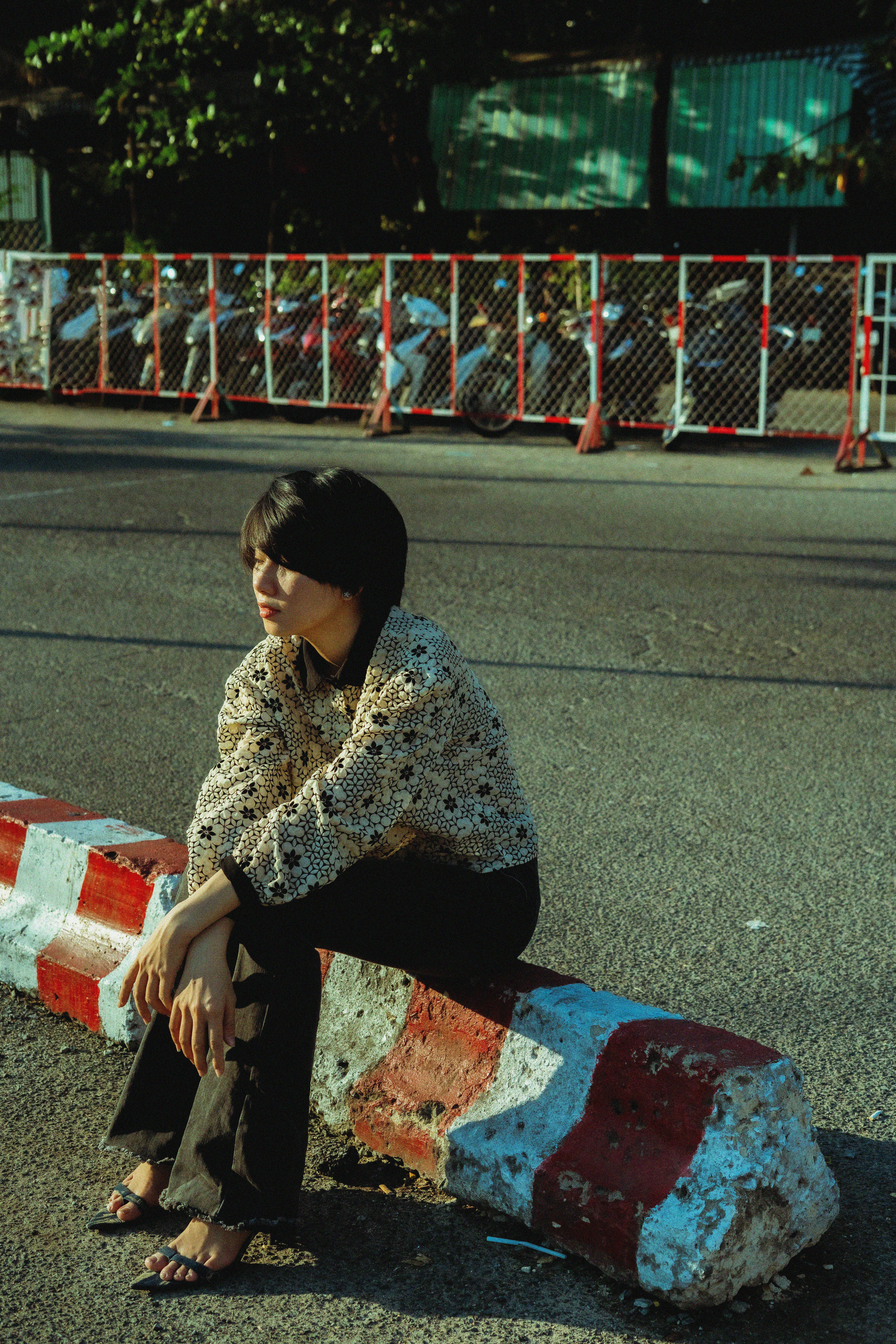 Stylish woman in patterned jacket poses on a city curb under sunlight, urban setting.
