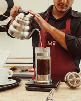 A barista pouring water over coffee grounds in a French press using a gooseneck kettle.