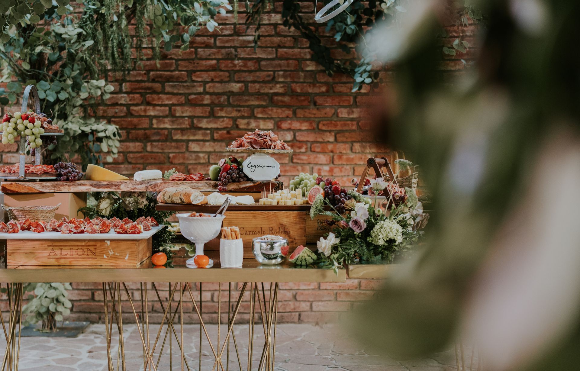 An elegantly arranged outdoor buffet table featuring a variety of snacks, fruits, and flowers against a brick wall.
