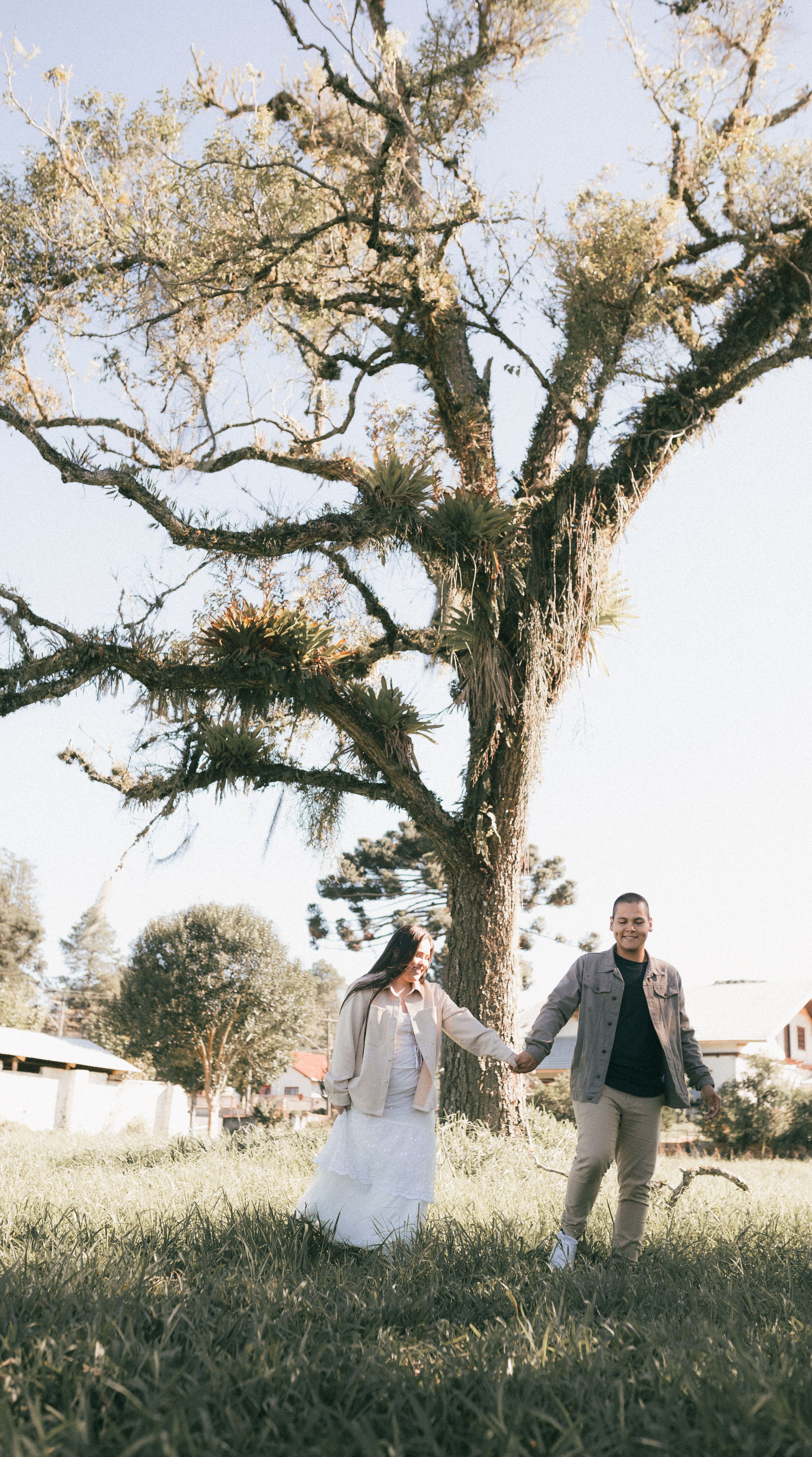 Smiling couple holding hands while walking through a sunny park with lush trees.