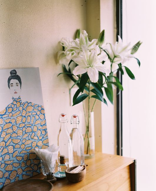 A modern dresser with a vase of lilies, artwork, and glass bottles against a beige wall.