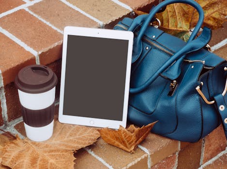 A stylish autumn still life with a coffee cup, tablet, and blue leather bag on brick steps adorned with fall leaves.