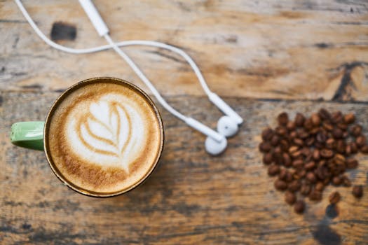 A cup of latte with artful foam next to earphones and coffee beans on a rustic wooden table.