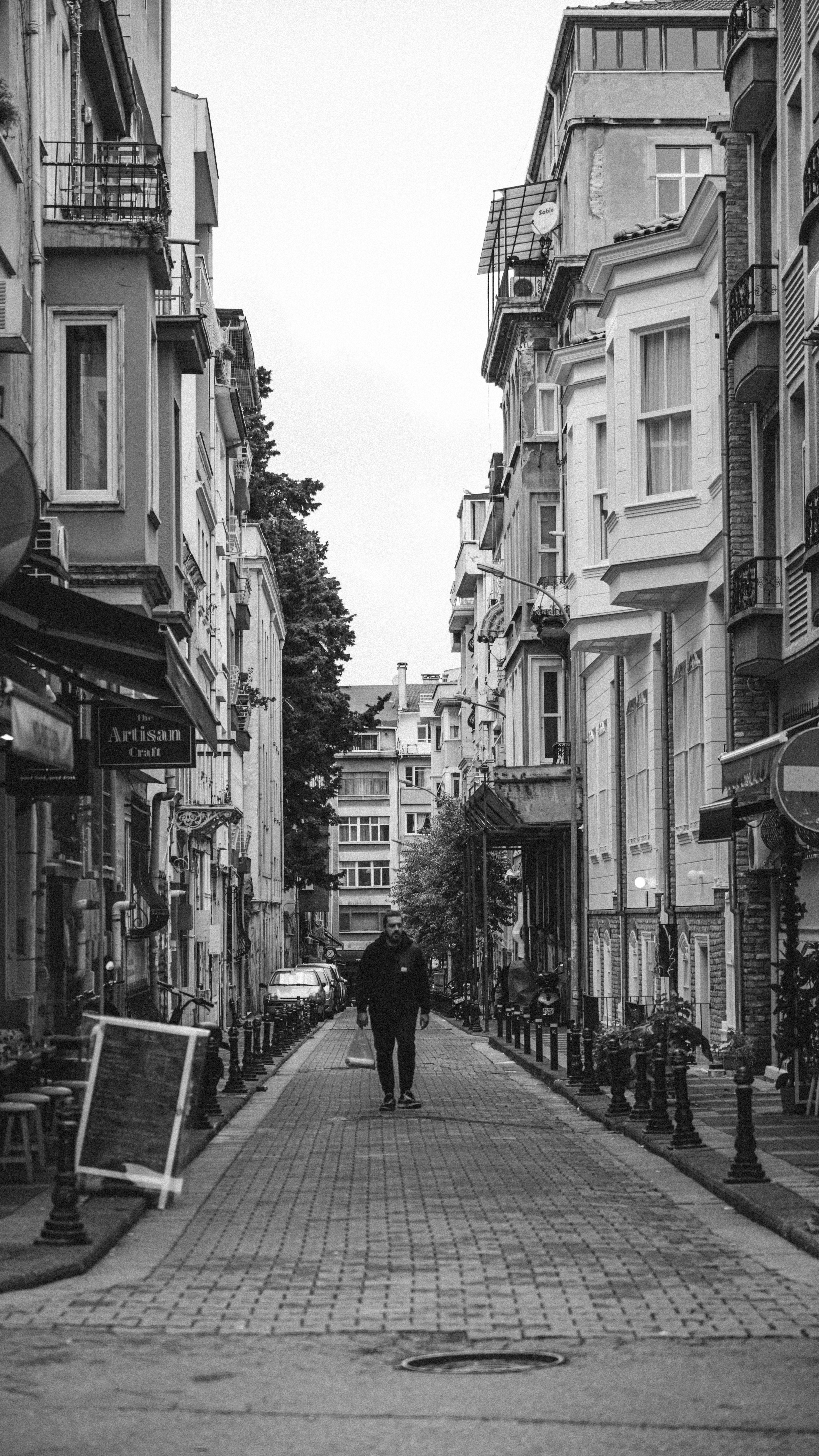 Black and white photo of a man walking on a narrow urban street lined with buildings.