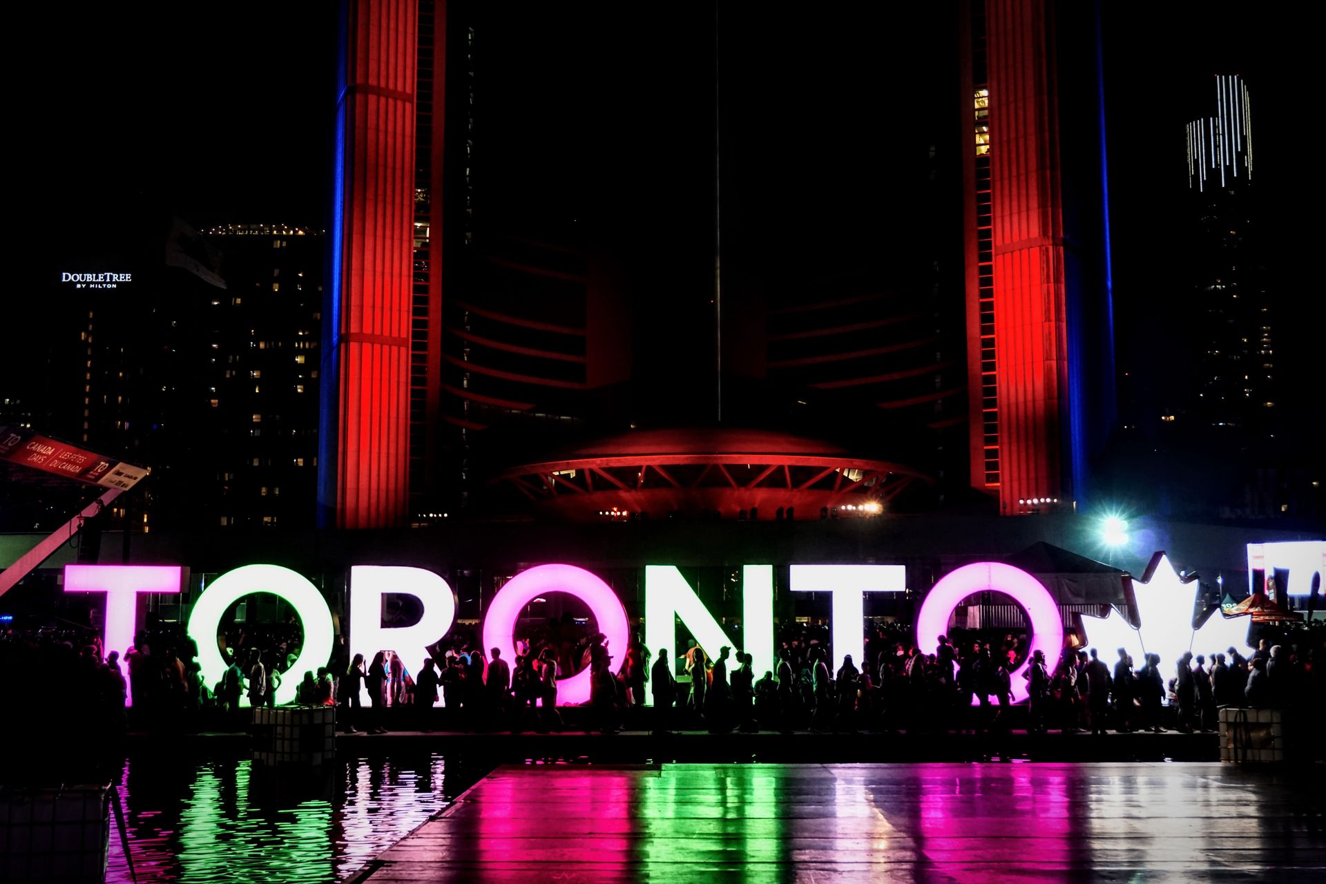 Vibrant Toronto sign glowing with colorful lights, featuring silhouetted crowd, iconic city symbol.