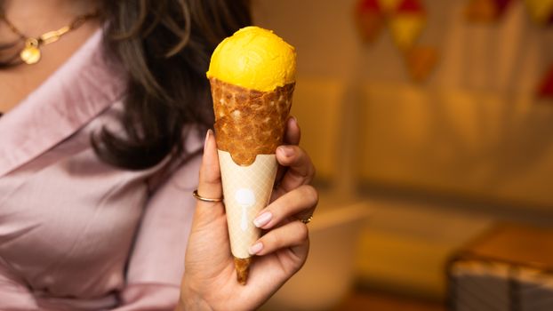 A woman holds a vibrant mango ice cream cone in a waffle cone, indoors.