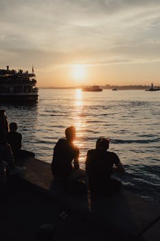 Silhouette of people relaxing at sunset by the Bosphorus in İstanbul, Türkiye, with ferries in the distance.