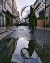城市街道的水坑倒映着建筑物的影子，人们撑着雨伞行走其间