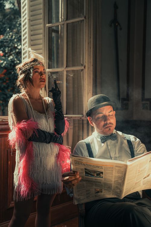 Free A man reading a newspaper and a woman smoking, both dressed in 1920s fashion, captured indoors with elegant lighting. Stock Photo