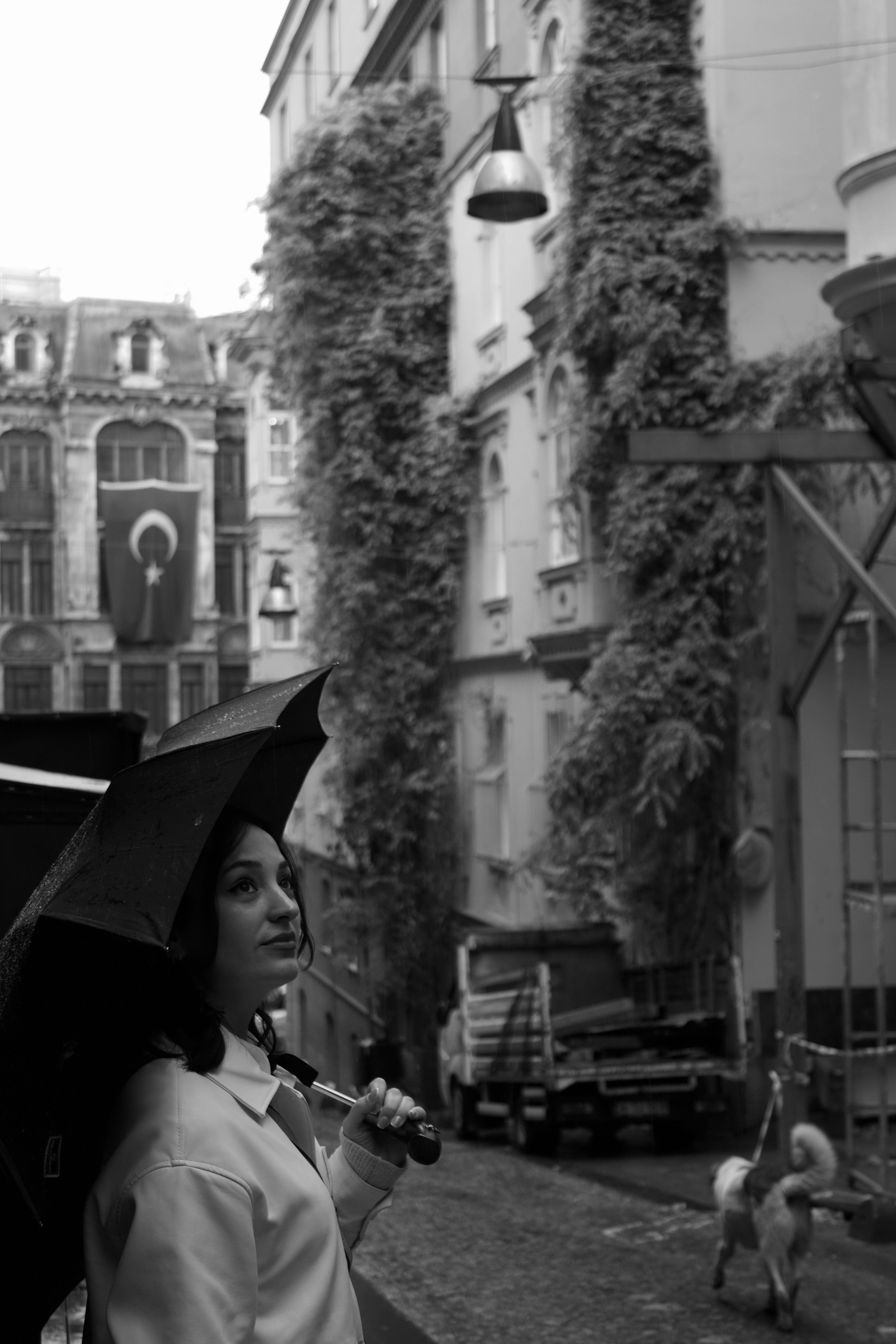 Black and white photo of a woman holding an umbrella gazing upwards in a city street.