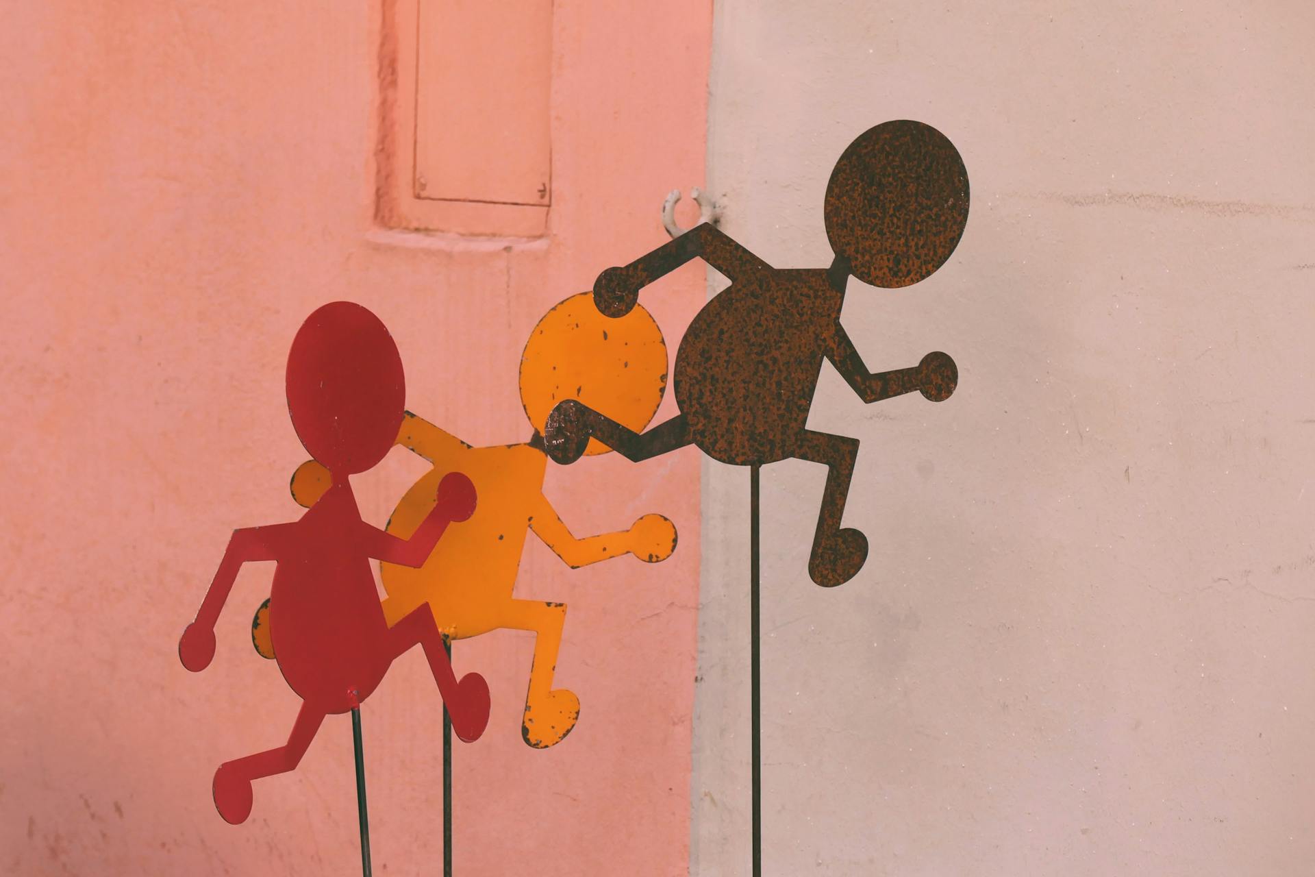Metal Anthropomorphic Figurines on Sticks Against a Pink Wall
