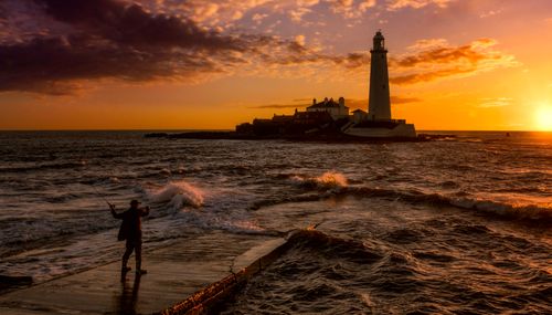Free A fisherman stands by St Mary's Lighthouse as waves crash during a vivid sunset. Stock Photo