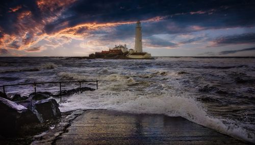 Free A towering lighthouse stands against a vibrant sunset over a stormy sea, creating a dramatic maritime scene. Stock Photo