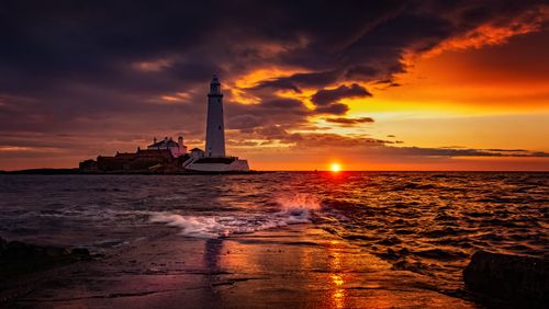 Free A tranquil sunset over the sea with a lighthouse and island in the background, reflecting vibrant colors in the calm waves. Stock Photo