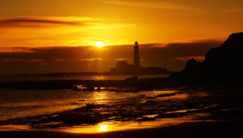 Free A striking silhouette of a lighthouse against a vibrant sunset sky on a tranquil beach. Stock Photo