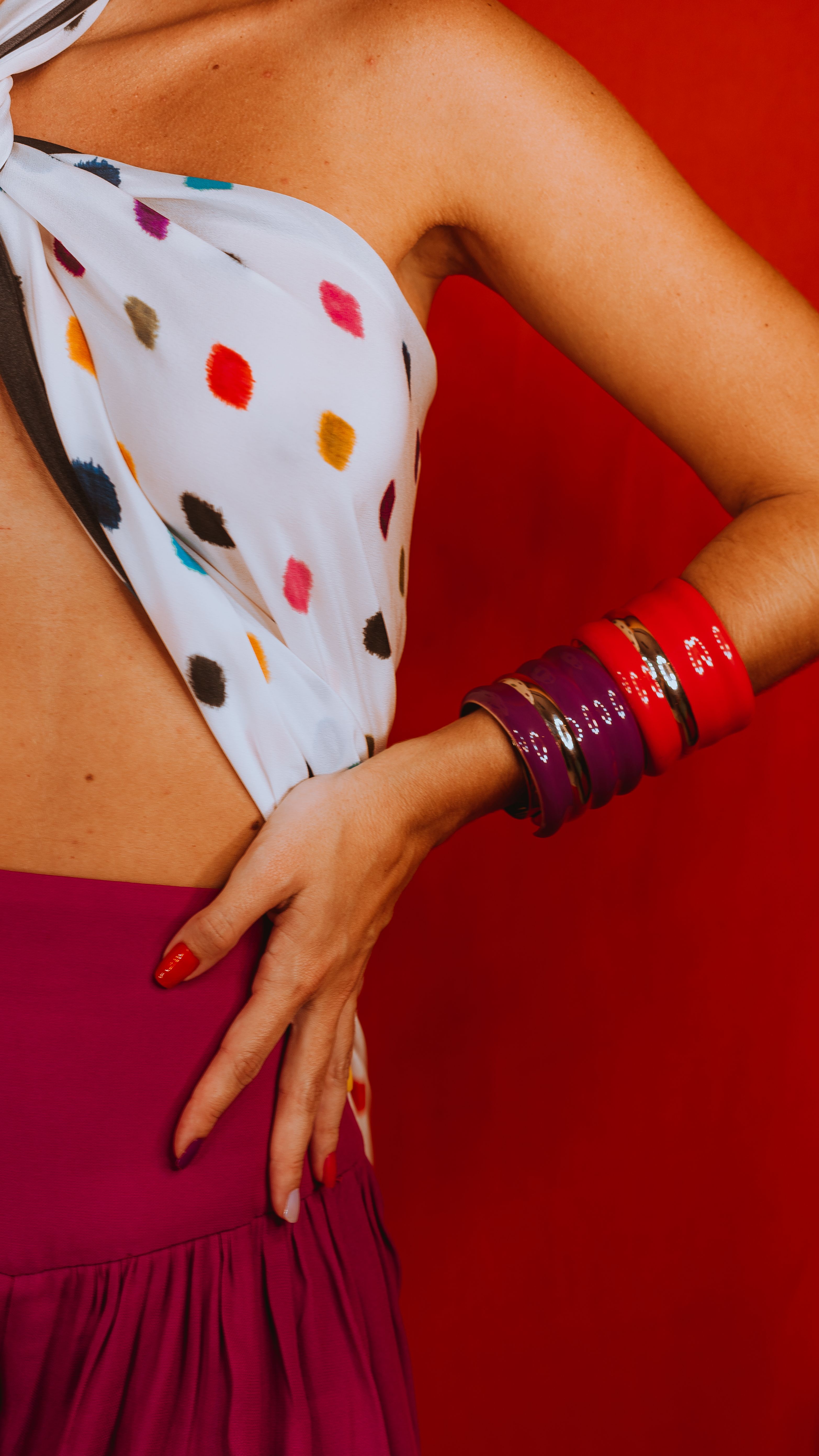 Mulher estilosa em top de bolinhas com pulseiras vibrantes e um fundo vermelho ousado.
