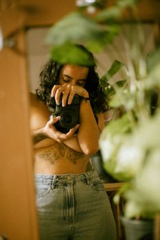 Young woman takes creative mirror selfie with camera, showcasing tattoos and indoor plants.