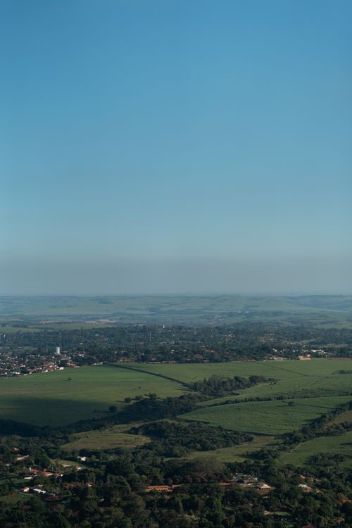 Free A breathtaking aerial view capturing expansive green fields and a small village under a clear blue sky. Stock Photo