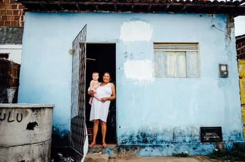 Free A cheerful woman holding a baby at the entrance of a blue rustic house in Natal, Brazil. Stock Photo