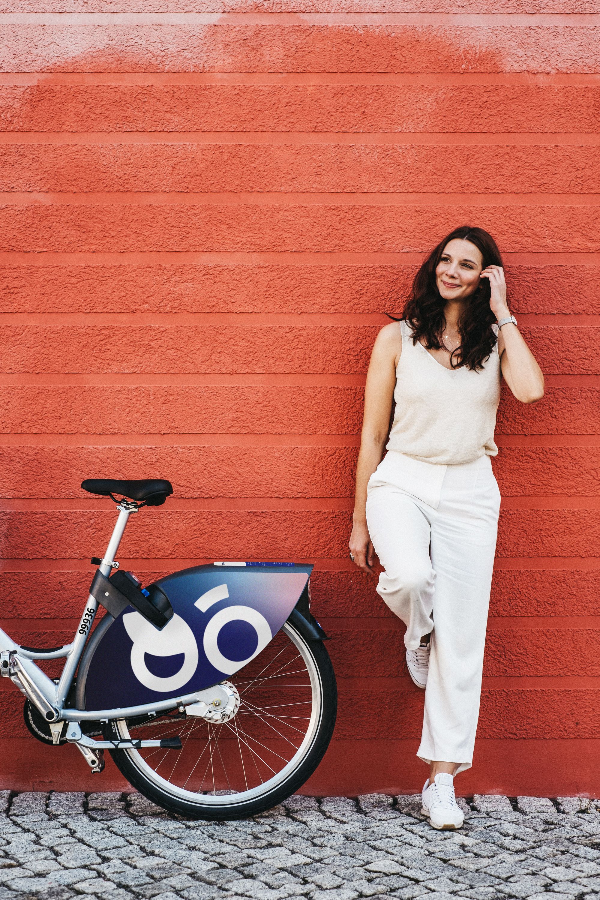 Smiling woman standing with a bicycle against a vibrant red wall, enjoying a casual moment.