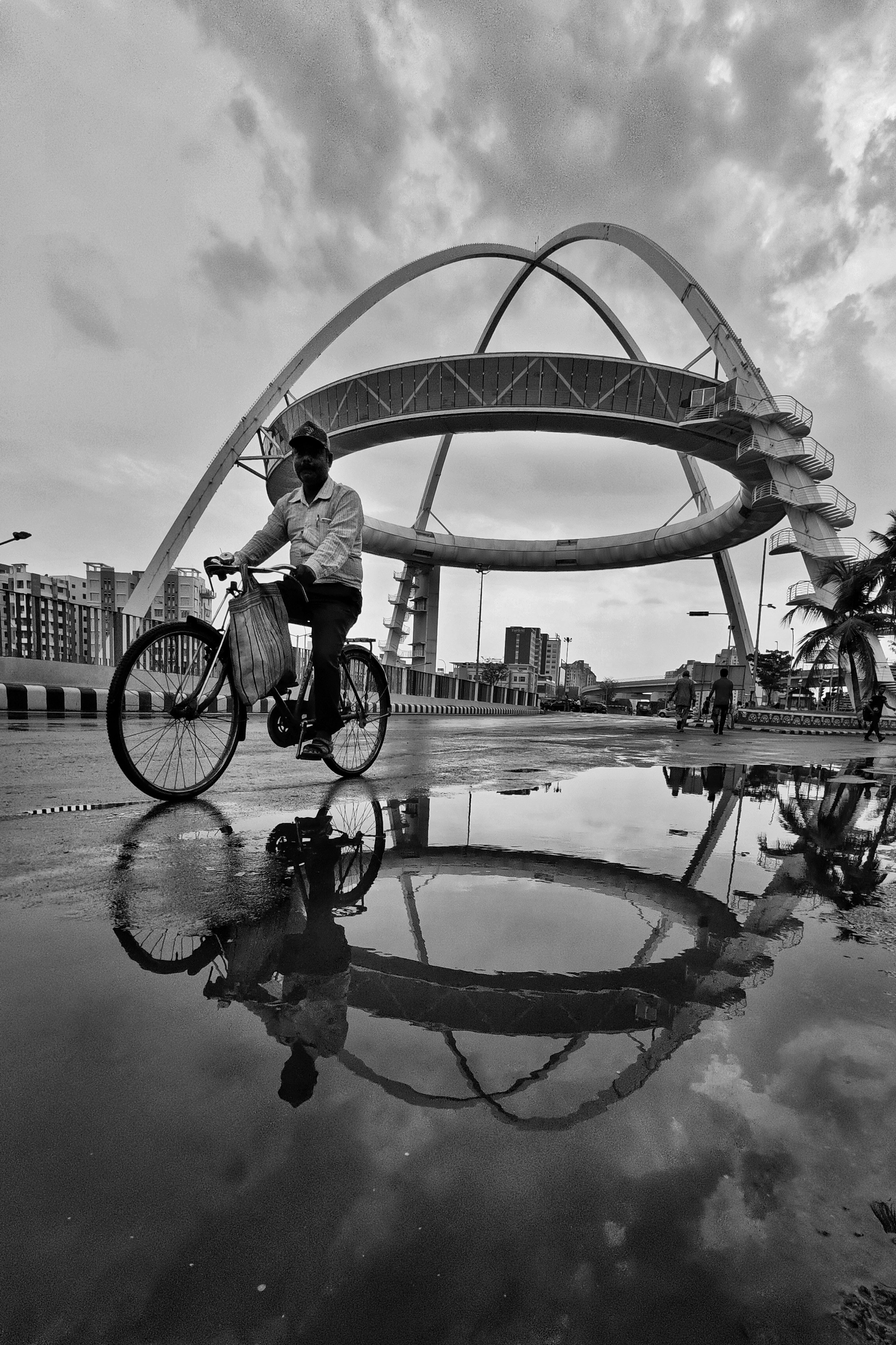 A cyclist rides by Biswa Bangla Gate with reflections in a rain puddle in Kolkata, India. Black and white capture.