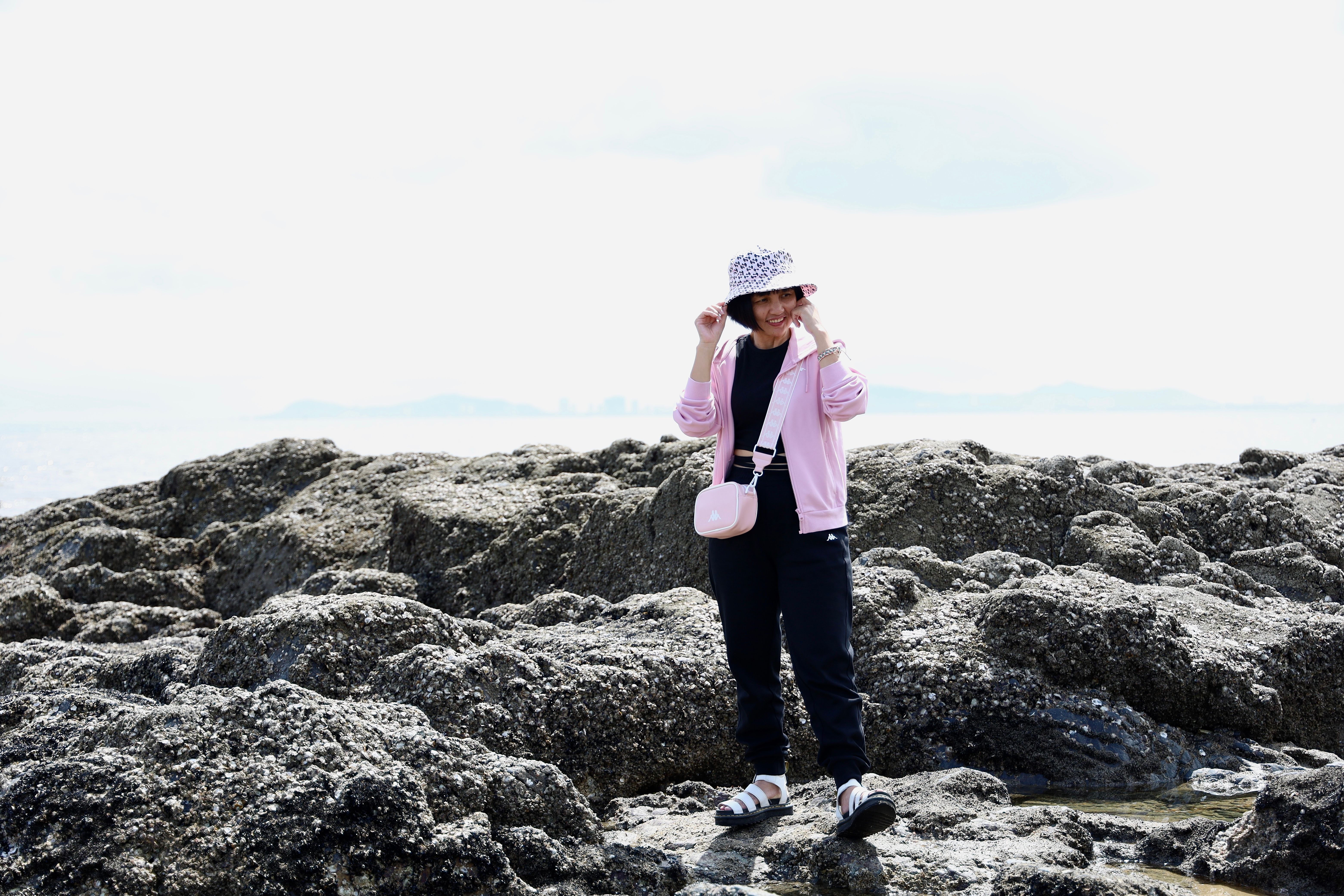 Asian woman standing on rocky shore in Vũng Tàu, Vietnam, wearing a pink jacket and summer hat.