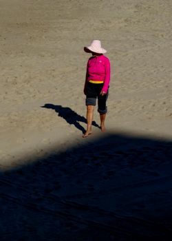 A woman in a pink top and summer hat walks barefoot on Alicante's sunny beach.