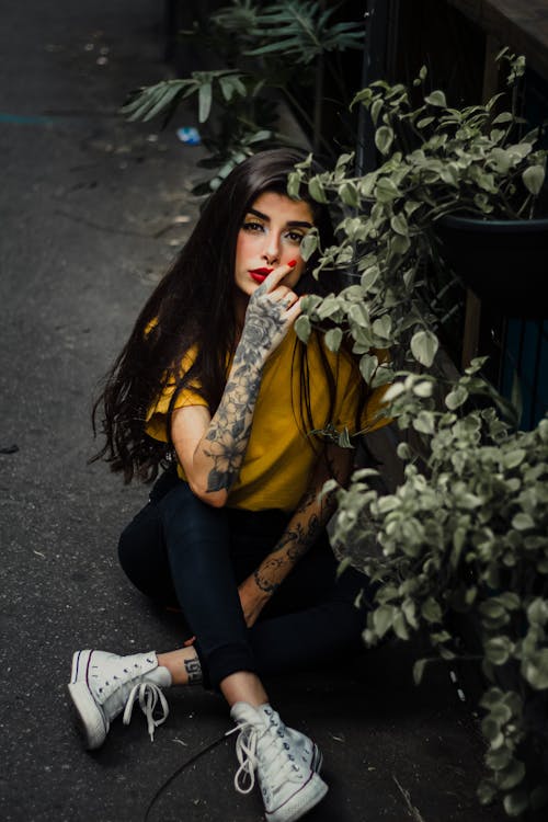 A woman with tattoos sitting on the ground next to plants