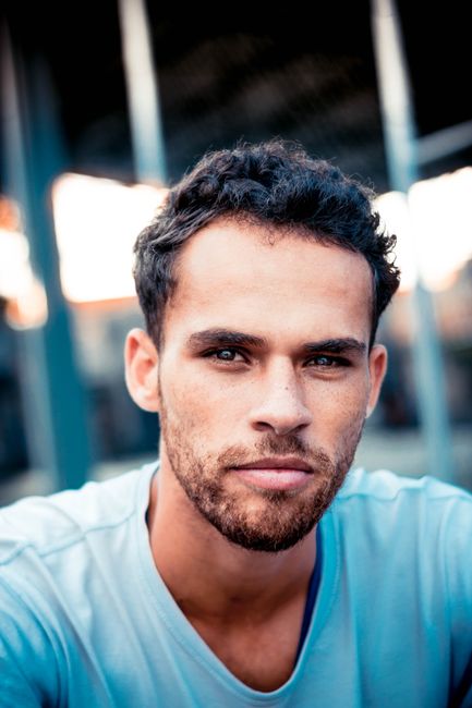 A close-up portrait of a young man with a confident expression, wearing a casual t-shirt outdoors.