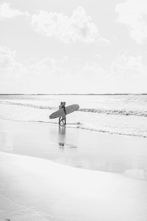 A black and white photo of a person walking on the beach with a surfboard