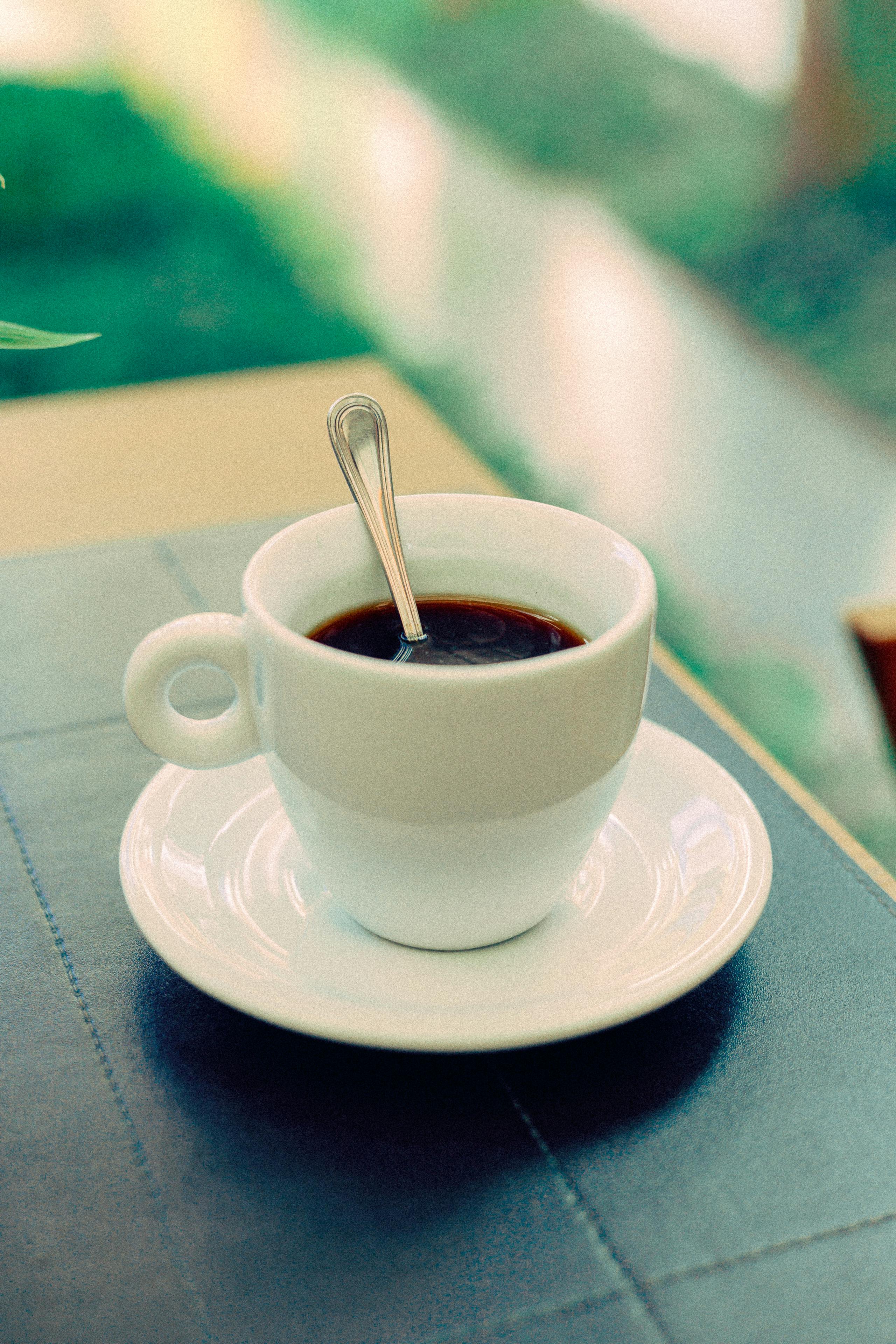 White cup of espresso with a spoon on saucer, set on a table in a café.