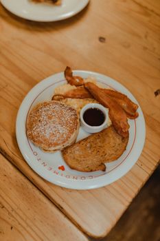 Top view of a savory breakfast platter with pancakes, bacon, and syrup on a wooden table.