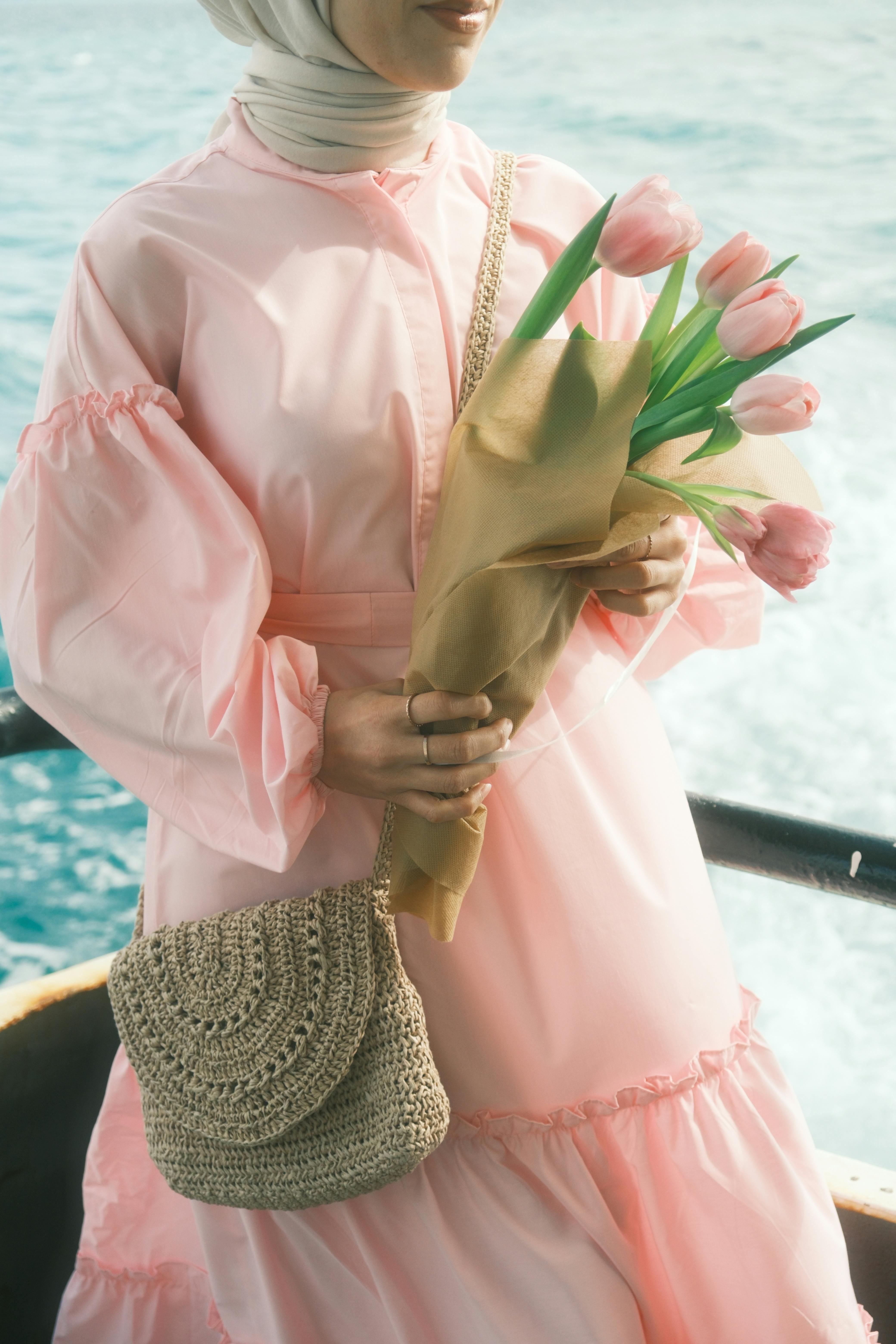 Elegant woman in a pink dress with hijab holding tulips, standing on a boat by the sea.