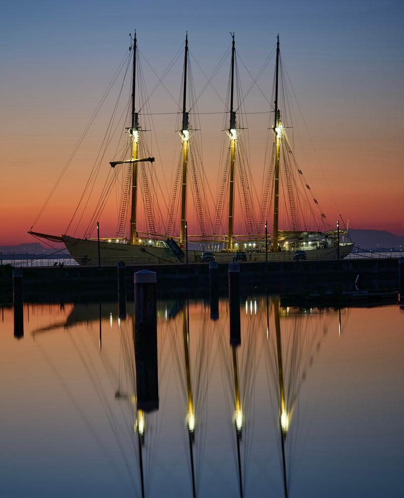 Sailing ship with tall masts moored on the shore at sunset reflected on calm water