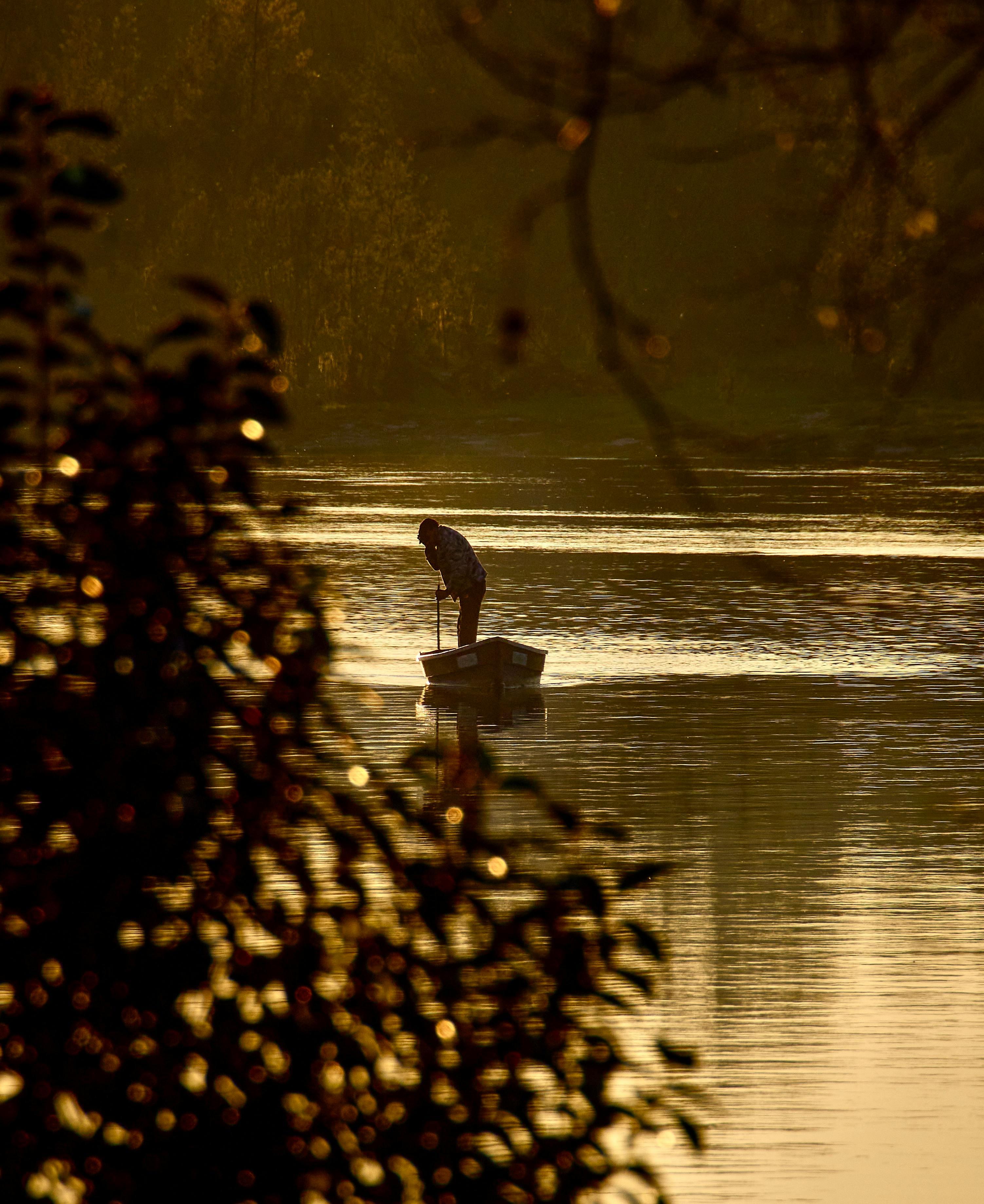 A lone figure stands in a small boat on a lake during sunset, with vibrant golden hues reflecting off the water.
