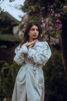 Stylish woman in a trench coat poses among pink blossoms, creating a serene and fashionable scene.