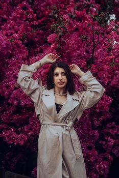 Elegant woman poses among vibrant pink flowers, showcasing chic fashion in a beige trench coat.