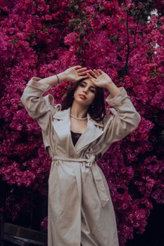 Fashionable woman in a beige trench coat posing gracefully in front of stunning pink bougainvillea flowers.