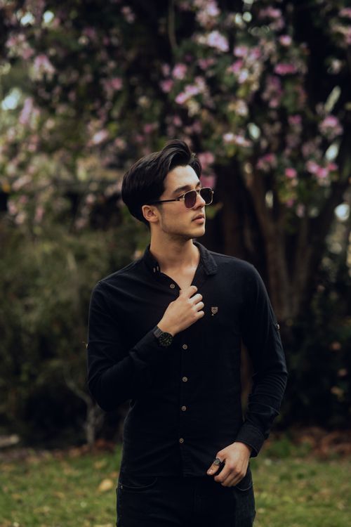 Free Young man in black shirt and sunglasses posing confidently outdoors with blooming trees. Stock Photo